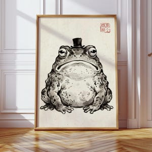 May include: A framed ink wash illustration of a toad wearing a small black top hat. The toad is rendered in shades of black and gray on a textured off-white background. A red square seal with Asian characters is visible in the top right corner.
