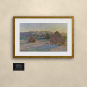 May include: A framed landscape painting depicting two large haystacks in a field under a soft sky, featuring muted tones of purple, gold, and green. Below the frame, a small black rectangular plaque displays white printed text.