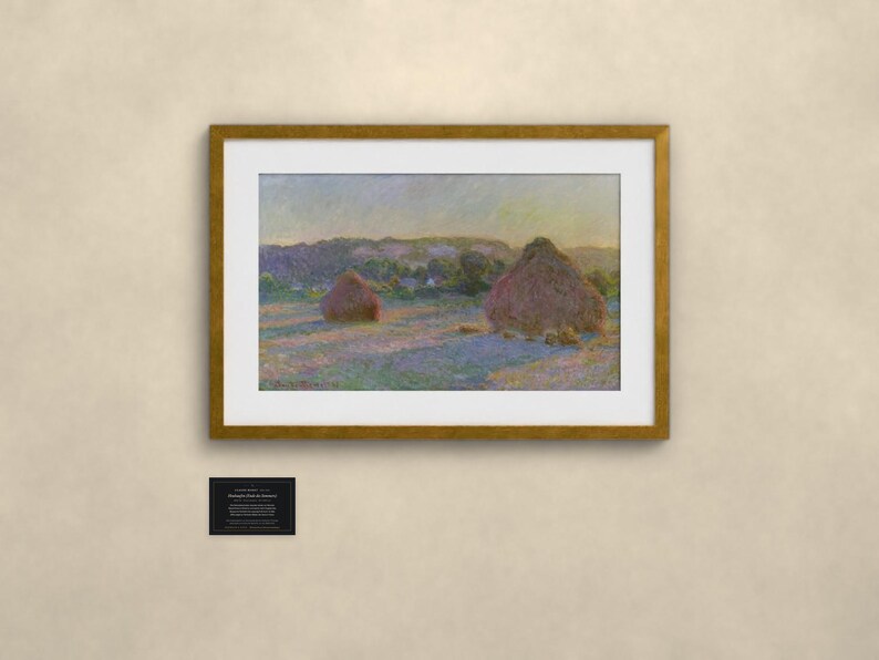 May include: A framed landscape painting depicting two large haystacks in a field under a soft sky, featuring muted tones of purple, gold, and green. Below the frame, a small black rectangular plaque displays white printed text.