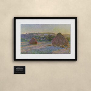 May include: A framed landscape painting depicting two large haystacks in a field under a soft, hazy sky. The artwork features muted tones of purple, yellow, and green. Below the frame, a small black plaque displays descriptive text.
