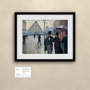 May include: A framed print of a painting depicting a rainy Paris street scene with people holding umbrellas. A couple walks in the foreground, while others traverse the cobblestone street near tall buildings. A small informational card is below.