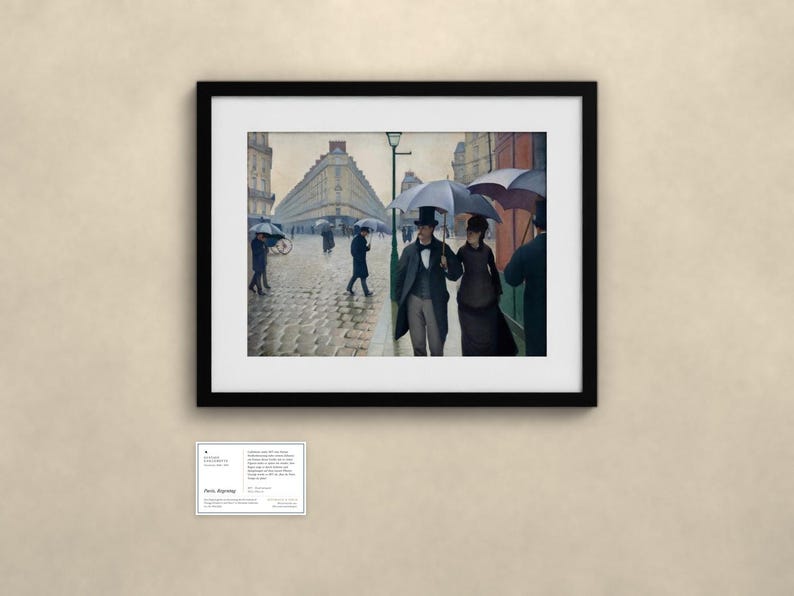 May include: A framed print of a painting depicting a rainy Paris street scene with people holding umbrellas. A couple walks in the foreground, while others traverse the cobblestone street near tall buildings. A small informational card is below.