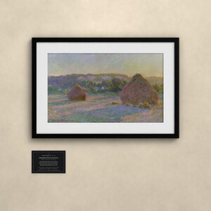 May include: A framed landscape painting depicting two haystacks in a field under a soft sky, featuring muted tones of purple, yellow, and green. Below the artwork, a small black rectangular plaque displays white text details.