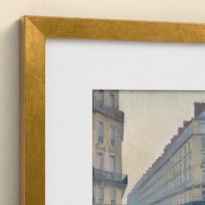 May include: A framed print featuring a painting of a city street with multi-storey buildings in muted tones of beige and blue. The artwork is enclosed in a wide, metallic gold-coloured frame with a clean white mat border surrounding the image.