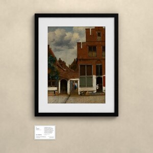 May include: A framed art print depicting a Dutch street scene with red brick buildings, a cobblestone path, and a cloudy sky. Below the frame, a small white informational card is displayed with the text The Little Street.