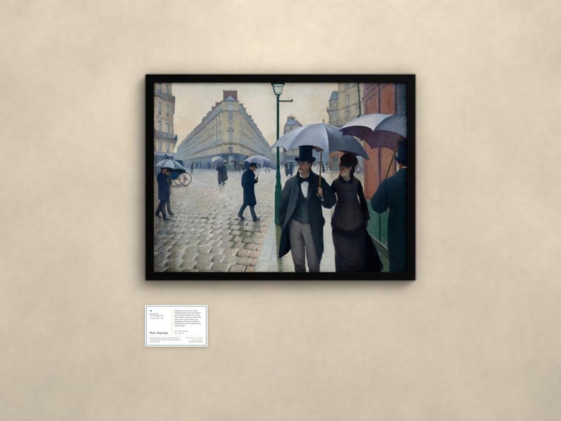 May include: A framed print of Gustave Caillebotte's Paris Street; Rainy Day, depicting figures with umbrellas walking on a cobblestone street in front of tall buildings. A small white informational plaque is mounted below the framed artwork.