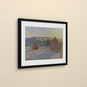 May include: A framed art print featuring an impressionist landscape painting of two haystacks in a field. The scene displays soft hues of purple, blue, and yellow under a pale sky. The artwork is enclosed in a simple, solid black rectangular frame.