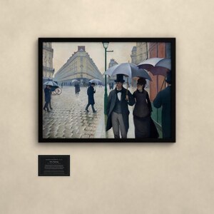 May include: A framed print of the painting Paris Street; Rainy Day by Gustave Caillebotte, depicting figures with umbrellas on a cobblestone street. Below the frame is a small black plaque with white text detailing the artwork information.