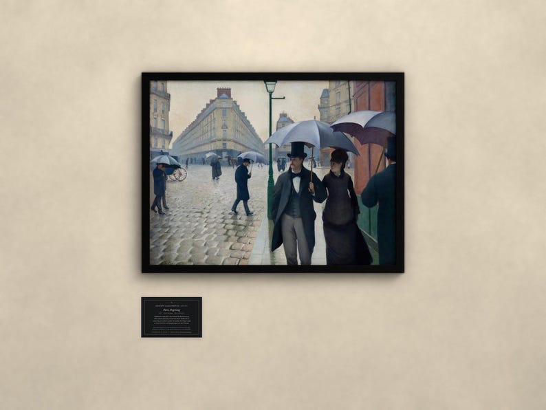 May include: A framed print of the painting Paris Street; Rainy Day by Gustave Caillebotte, depicting figures with umbrellas on a cobblestone street. Below the frame is a small black plaque with white text detailing the artwork information.