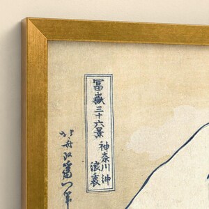 May include: A framed print featuring a cream-colored background with dark blue Japanese calligraphy and a partial outline of a mountain. The artwork is enclosed in a metallic gold frame with a textured finish, highlighting the contrast between the paper.