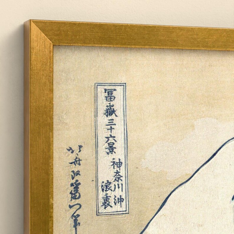 May include: A framed print featuring a cream-colored background with dark blue Japanese calligraphy and a partial outline of a mountain. The artwork is enclosed in a metallic gold frame with a textured finish, highlighting the contrast between the paper.