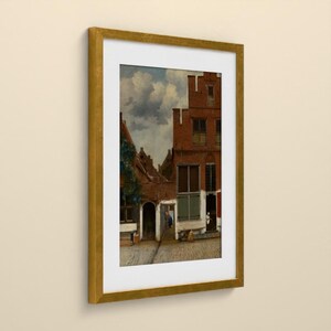 May include: A framed art print depicting a street scene with red brick buildings, a cobblestone path, and a cloudy sky. The artwork is enclosed in a gold-colored frame with a white mat, showcasing architectural details and figures in a courtyard.