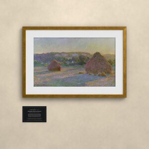 May include: A framed art print featuring an impressionist landscape painting of two haystacks in a field at sunset. The scene displays soft purple and gold light across the grass, with a small black informational plaque mounted below the frame.