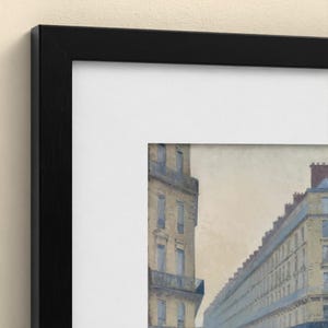 May include: A framed print featuring a painting of a city street scene with tall, multi-storey buildings in muted beige and blue tones. The artwork is encased in a wide white mount and a thin, black wooden frame with a visible wood grain texture.