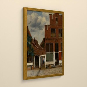 Op de afbeelding: Een ingelijst olieverfschilderij met een Nederlands straatbeeld met rode bakstenen gebouwen, groene luiken en een kasseienpad onder een bewolkte lucht. Het kunstwerk zit in een gouden lijst en toont architectonische details en figuren.