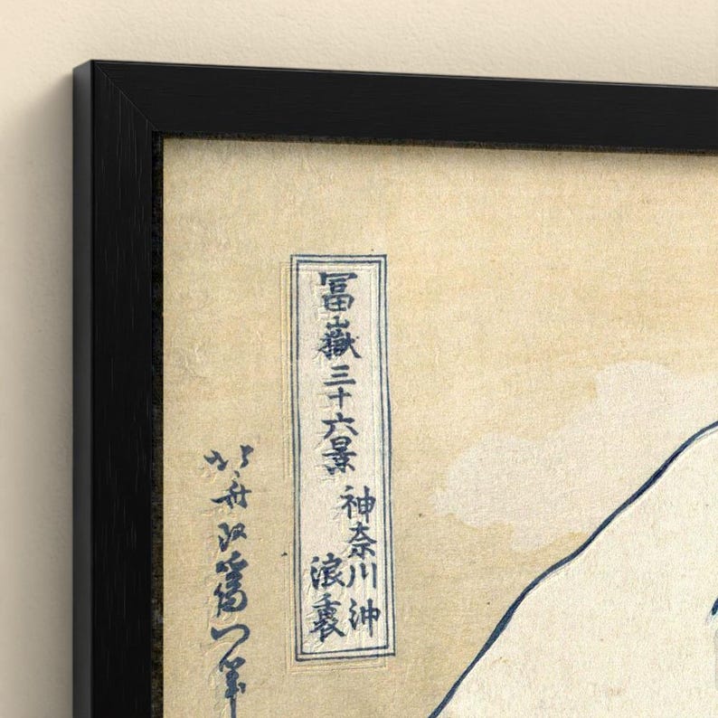 May include: A framed art print featuring a cream-colored background with dark blue Japanese calligraphy and a partial outline of a mountain. The artwork is enclosed in a simple, thin black frame that borders the entire rectangular composition.