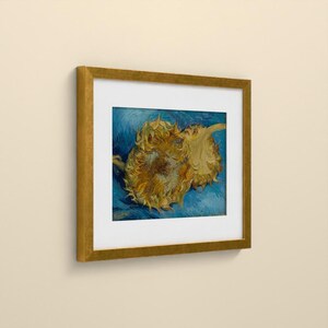 May include: A framed art print featuring a painting of two yellow sunflowers against a textured blue background. The artwork is enclosed in a white mat and a metallic gold frame, displaying a signature in the bottom left corner.