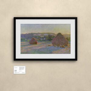 May include: A framed landscape painting featuring two large haystacks in a field under a soft sky. The artwork displays muted tones of yellow, purple, and green. Below the frame, a small white informational placard contains printed text.