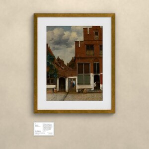 May include: A framed art print depicting a Dutch cityscape with red brick buildings, a cobblestone street, and a cloudy sky. The artwork is enclosed in a gold-colored frame and is accompanied by a small white informational label placed below it.