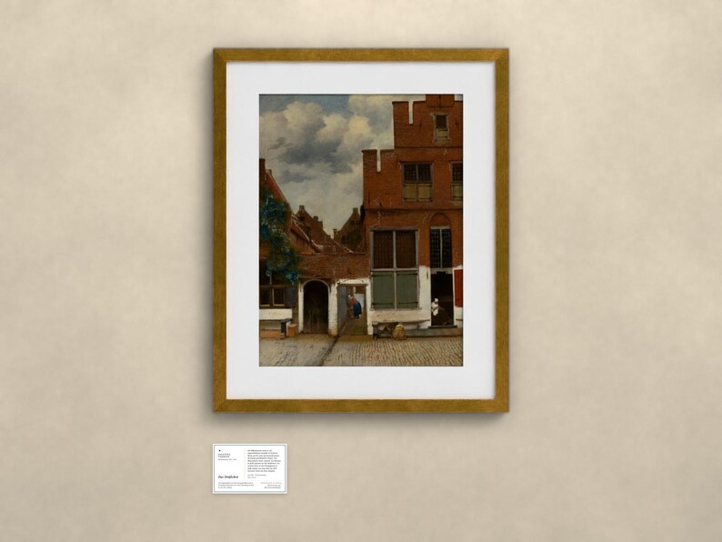 May include: A framed art print depicting a Dutch cityscape with red brick buildings, a cobblestone street, and a cloudy sky. The artwork is enclosed in a gold-colored frame and is accompanied by a small white informational label placed below it.