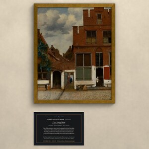 Op de afbeelding: Een ingelijste kunstdruk van Johannes Vermeer's schilderij Het Straatje, met een rood bakstenen huis met trapgevel, een geplaveide binnenplaats en figuren, vergezeld van een zwart informatiebordje met witte tekst.