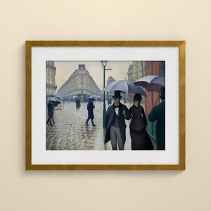 May include: A framed print of a painting depicting a rainy Parisian street scene with cobblestones, tall buildings, and people walking with umbrellas. A couple in the foreground wears dark coats and hats, while others walk in the distance.