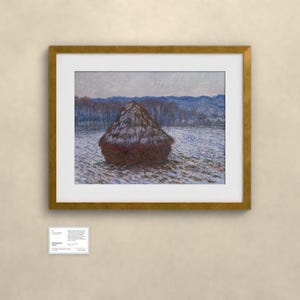 May include: A framed landscape painting featuring a dark brown haystack in a snow-covered field under a pale sky, with blue-toned hills in the distance. A small white informational placard is positioned below the gold-toned frame.