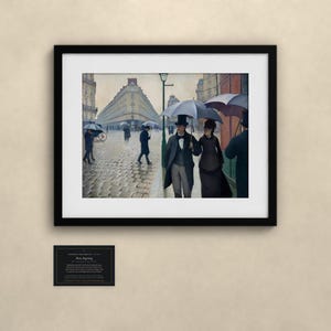 May include: A framed art print depicting a rainy Paris street scene with people holding umbrellas, including a couple in the foreground. The painting features cobblestone streets, tall buildings, and a street lamp. A small informational plaque is below the frame.