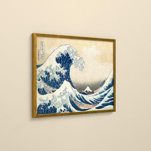 May include: A framed print of The Great Wave off Kanagawa featuring a large, cresting blue wave with white foam, small boats, and a distant Mount Fuji against a beige background. The artwork includes vertical Japanese text on the left side.