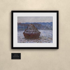 May include: A framed print of a landscape painting featuring a dark, conical haystack centered in a snow-covered field. The background shows blue-toned trees and hills under a pale sky. A small black plaque with white text is mounted below the frame.