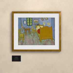 May include: A framed print of Vincent van Gogh's Bedroom in Arles painting, featuring a yellow bed, two yellow chairs, a wooden table, and blue walls, enclosed in a gold-colored frame with a small black informational plaque below it.