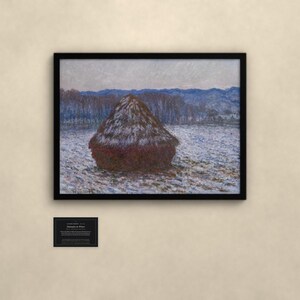 May include: A framed landscape painting depicting a large, dark brown haystack centered in a snow-covered field. In the background, a line of trees and distant blue mountains are visible under a pale sky. A small informational plaque is below the frame.