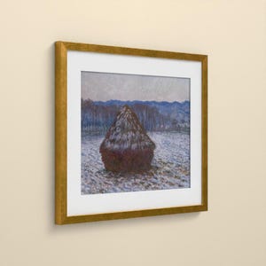 May include: A framed print of an impressionist painting featuring a dark brown haystack centered in a snowy field. The background displays blue-toned hills under a pale sky. The artwork is enclosed in a square, metallic gold frame with a white mat.