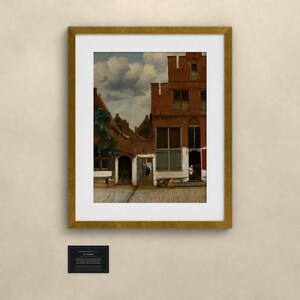 May include: A framed print of a painting depicting a Dutch street scene with red brick buildings, a cobblestone path, and a cloudy sky. The artwork is enclosed in a gold-toned frame with a white mat. A small black informational plaque is below the frame.