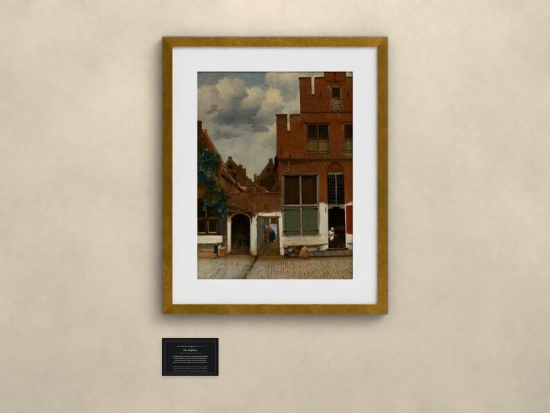 May include: A framed print of a painting depicting a Dutch street scene with red brick buildings, a cobblestone path, and a cloudy sky. The artwork is enclosed in a gold-toned frame with a white mat. A small black informational plaque is below the frame.