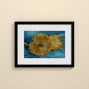 May include: A framed art print featuring a painting of two withered sunflowers with golden-brown petals resting against a textured, vibrant blue background. The artwork is enclosed in a simple black frame with a wide white mat border.
