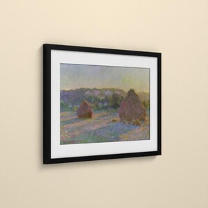 May include: A framed art print featuring an impressionist landscape painting of two haystacks in a field. The scene displays soft hues of purple, blue, and yellow under a pale sky. The artwork is enclosed in a simple, solid black rectangular frame.
