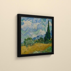 May include: A framed square art print featuring a landscape painting with a golden wheat field, green trees, and a tall dark cypress tree under a swirling blue and white sky. The artwork is enclosed in a simple black frame.