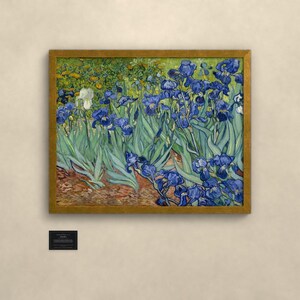 May include: A framed painting of blue irises with green leaves and a single white iris, set against a background of yellow flowers and brown earth. The artwork is enclosed in a gold-toned frame and features a small black plaque below it.