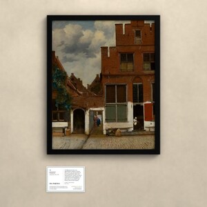 May include: A framed print of a painting depicting a Dutch courtyard with red brick buildings, a cobblestone ground, and a cloudy sky. A small white information card is positioned below the frame, containing text about the artwork.