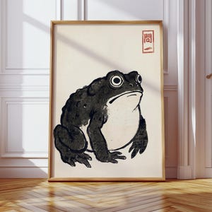 May include: A framed wall art print featuring a black ink illustration of a toad with a light-colored belly on a cream background. In the upper right corner, there is a red rectangular stamp containing the Japanese characters 問一.