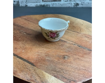 May include: A small white porcelain teacup featuring a delicate pink rose floral pattern on the front and a thin gold-coloured rim along the top edge. The teacup has a single handle on the right side and a smooth, glossy finish throughout.