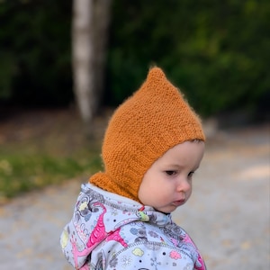 Könnte beinhalten: Eine gestrickte senfgelbe Zipfelmütze mit geripptem Rand, getragen über einer hellgrauen Kapuzenjacke mit einem bunten Grafikdruck aus rosa Dinosauriern, gelben Wolken und schwarzen Wirbelmustern auf weißem Hintergrund.