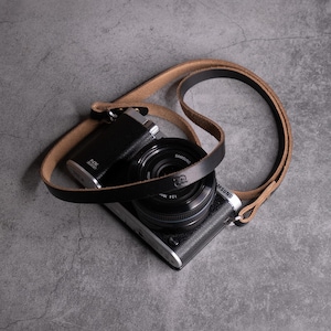 May include: A black and silver Samsung NX3000 digital camera with a black lens, featuring a dual-toned camera strap made of smooth black leather on the exterior and a natural tan suede-like texture on the interior, attached to the camera body.