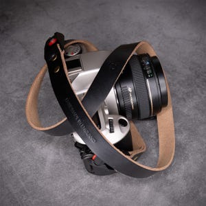 May include: A black leather camera strap with a natural tan underside, featuring gold-tone metal rivets and red accents. The strap is embossed with the text HANDMADE IN ENGLAND and is attached to a silver and black camera body with a black lens.