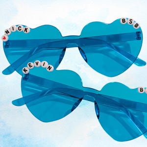 Puede incluir: Dos pares de gafas de sol en forma de corazón en un tono azul vibrante. Cada par presenta detalles de cuentas que deletrean "NICK", "KEVIN" y "BSB". Las gafas tienen montura y lentes transparentes, perfectas para un look divertido y con estilo.