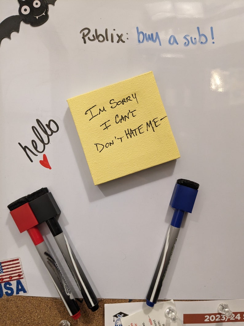 May include: A white dry-erase board with a yellow sticky note that says "I'm sorry I can't Don't hate me-". There are two black and white dry-erase markers with red and blue caps, a small American flag, and a bat drawing on the board.