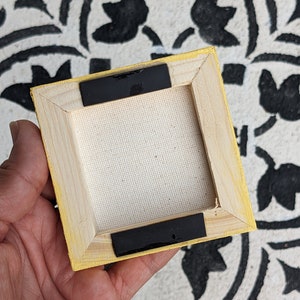 May include: The back of a small, yellow-painted wooden canvas frame with black tape on the edges. The canvas is white and the frame is made of wood.