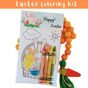 Pode incluir: Um kit de colorir de Páscoa com um cartão branco que apresenta um coelho, um pintinho e uma cesta. O cartão tem uma faixa de "Feliz Páscoa" e um sol com um rosto sorridente. O kit inclui cinco lápis de cera e uma conta de madeira em forma de cenoura com um tassel verde.