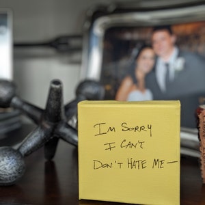 May include: A yellow sticky note with the handwritten message "I'm sorry I can't Don't hate me" on a wooden surface. A blurry photo of a couple is in the background.